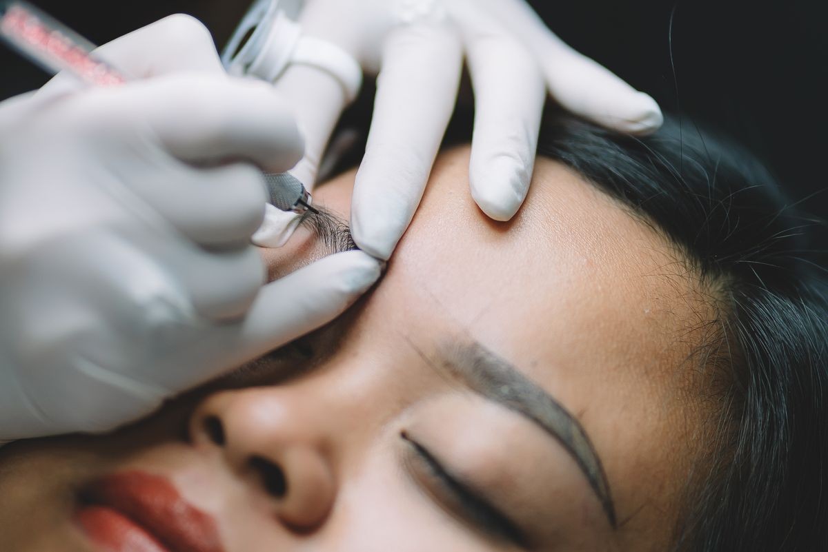 Beautiful asian woman having professional eyebrow tattoo in beauty salon. Cosmetologist applying permanent make up on eyebrows.